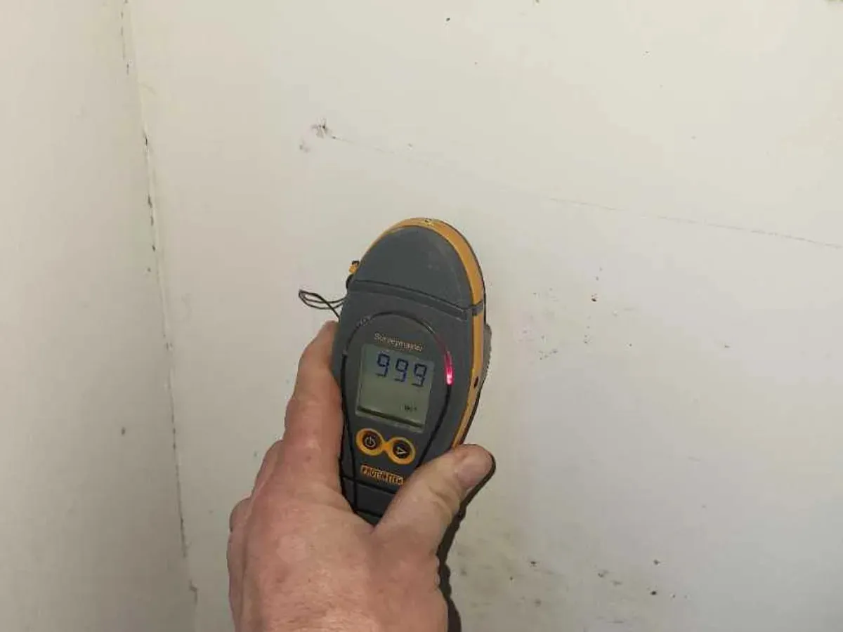 A moisture meter held against an interior wall displays a high reading