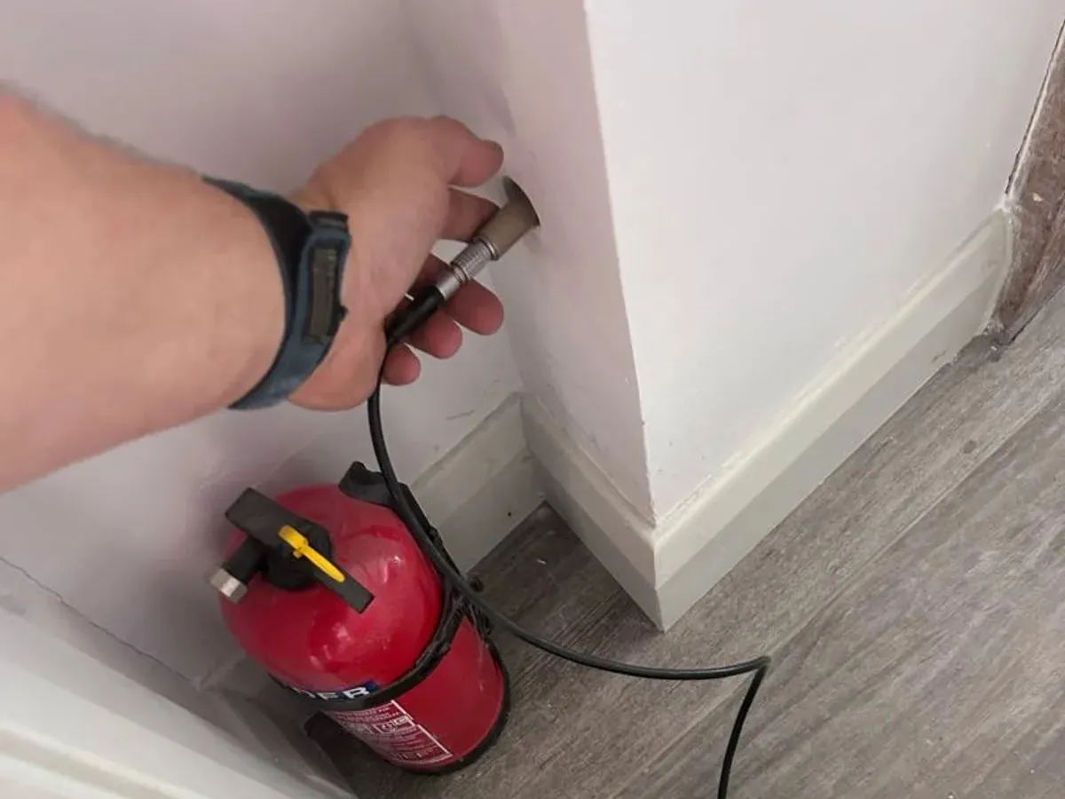 An engineer holding a tracer gas probe against white kitchen boxing above the skirting board
