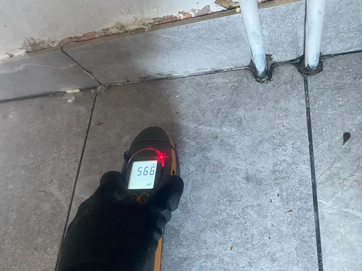 A gloved hand holds a moisture meter against grey floor tiles beside wall pipework